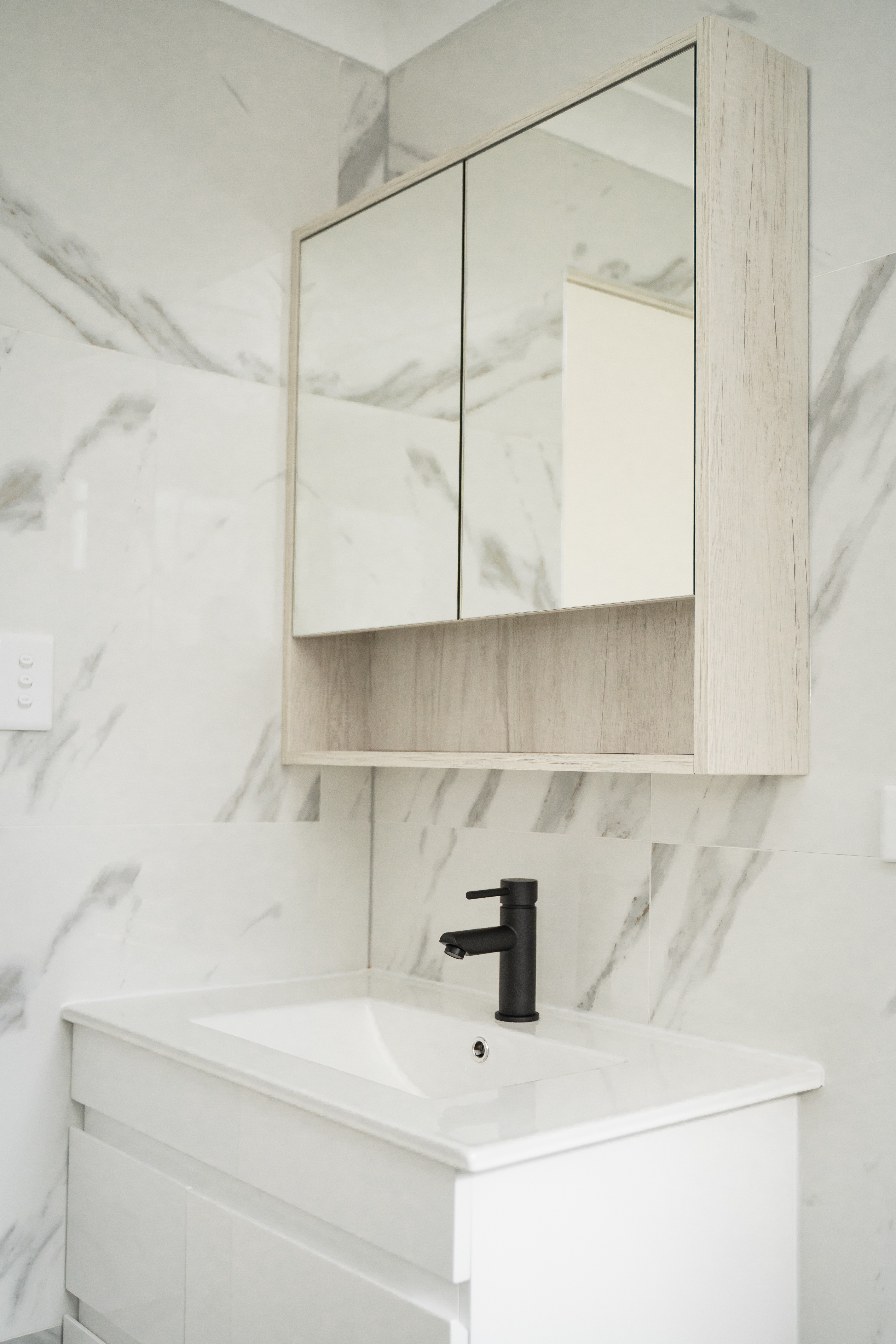 Vanity — three-door timber mirror cabinet above white basin, marble wall tiles