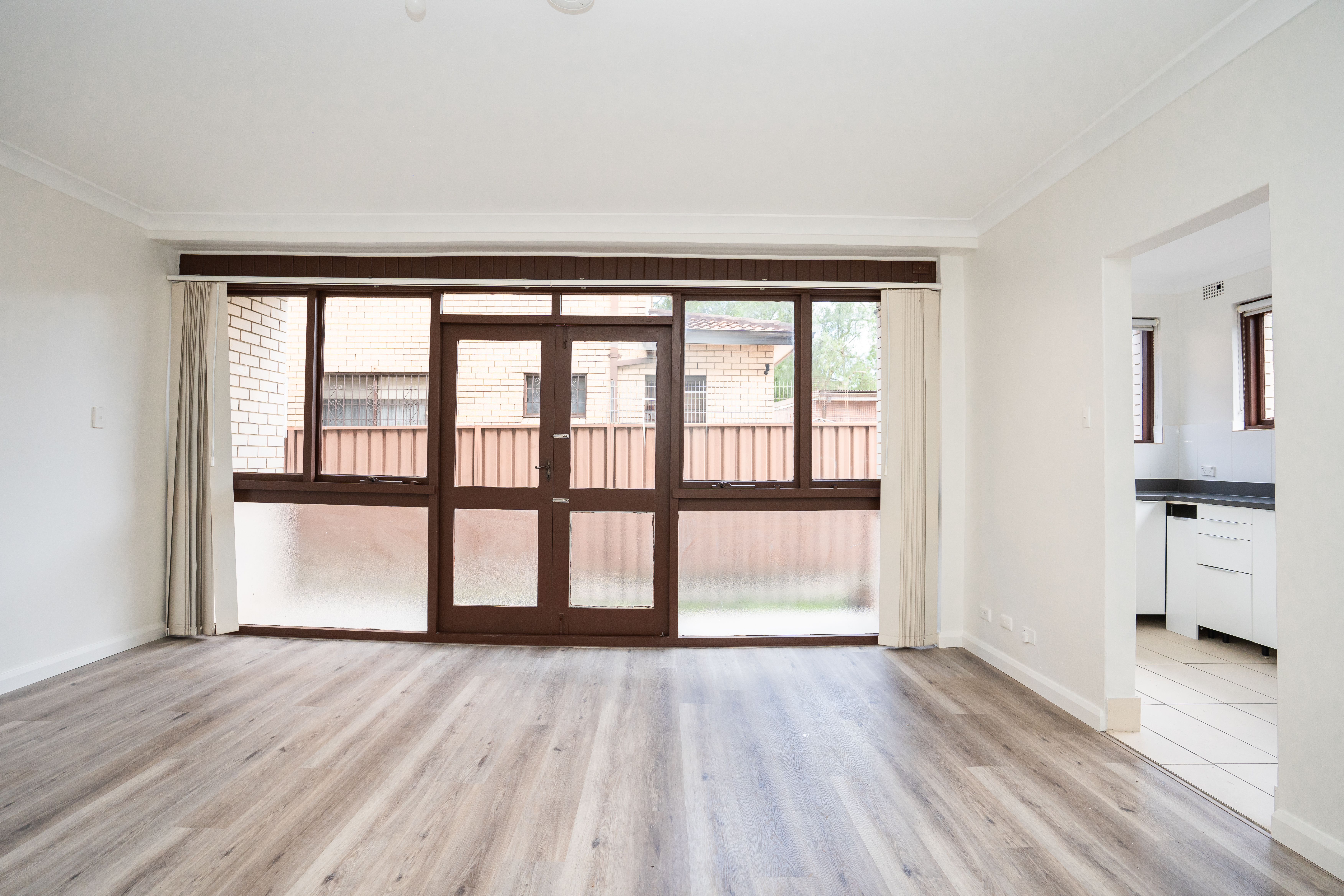 Living area — wide view with timber flooring, curtains, and kitchen pass-through