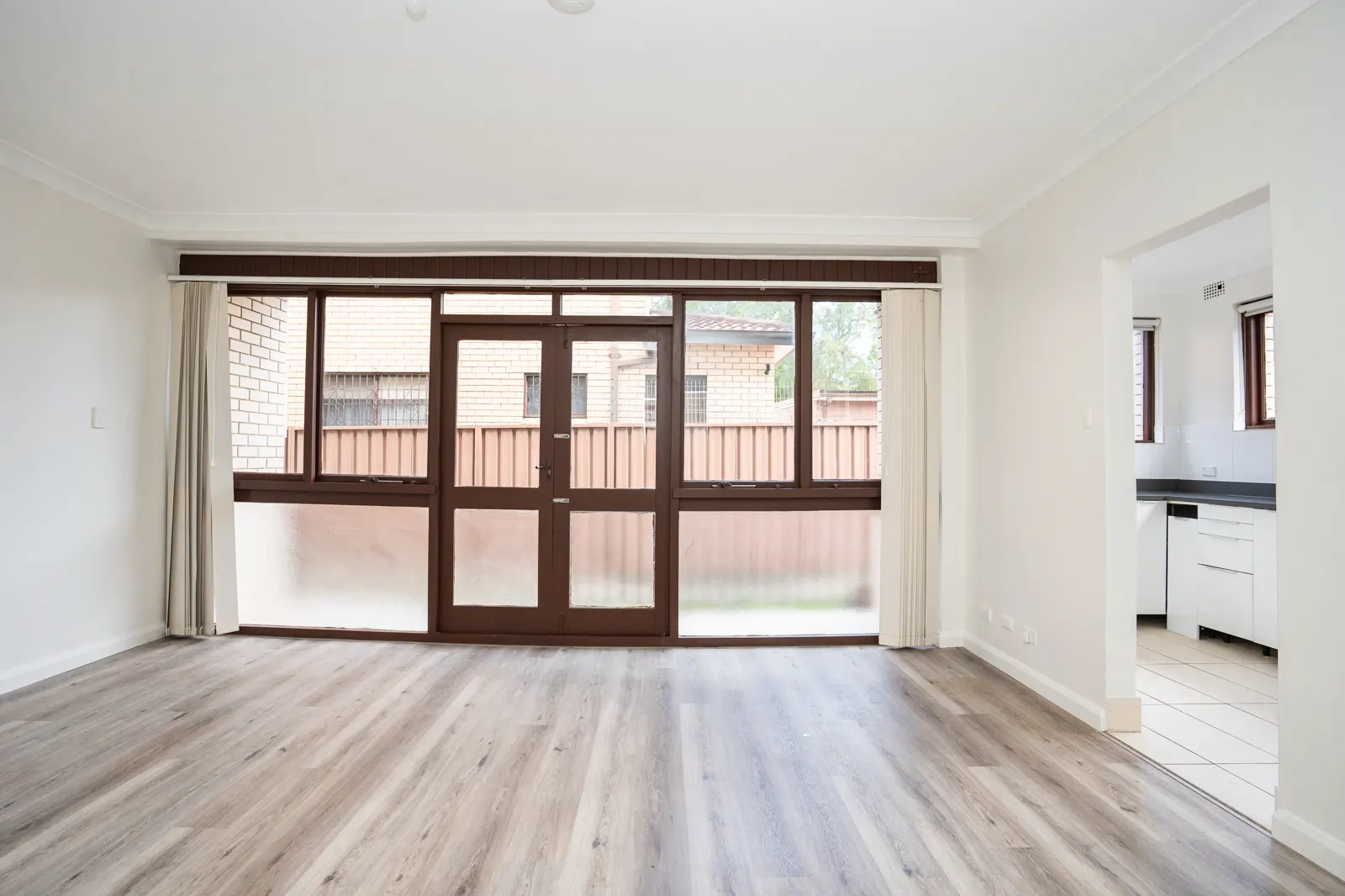 Living area — wide view with timber flooring, curtains, and kitchen pass-through