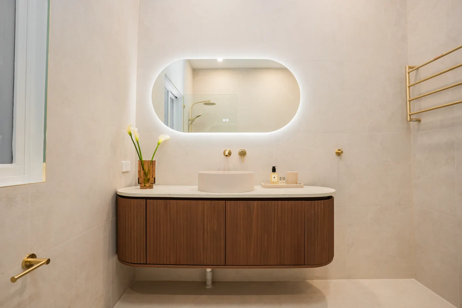 Floating walnut vanity with stone top, backlit oval mirror and wall-mounted gold tapware on porcelain tiles