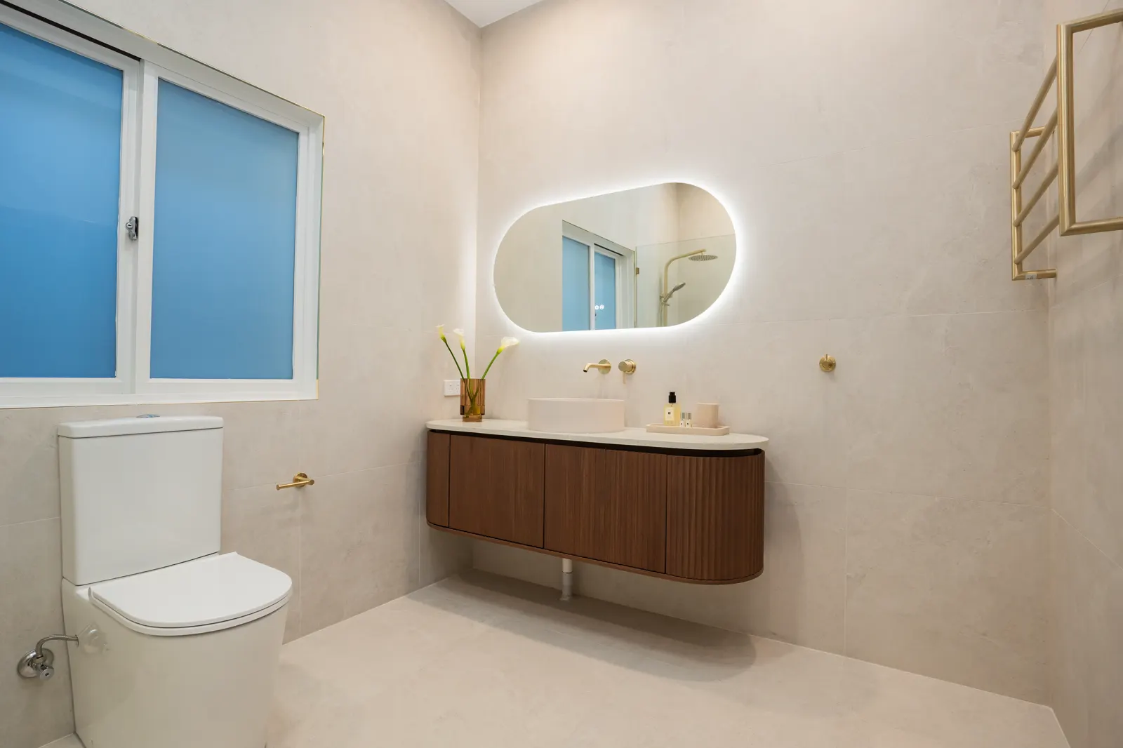 Fluted walnut vanity and backlit mirror against large-format wall tiles with brushed gold towel rail
